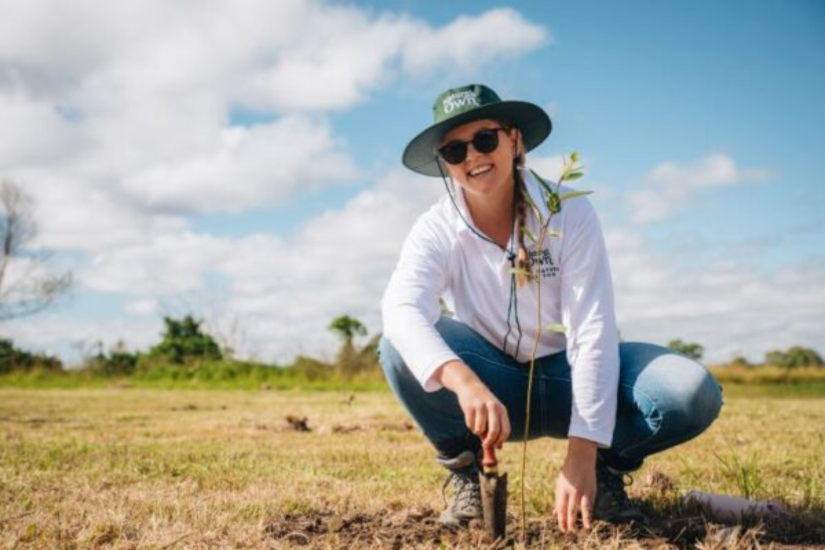 A person kneeling on the ground, planting a young tree in a grassy field, wearing a hat and sunglasses.